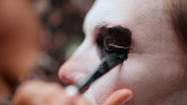 Close-up of makeup artist applying black eye shadow around closed eye for Day of the Dead or Halloween costume. Brush strokes create dramatic dark circles on white face paint base.