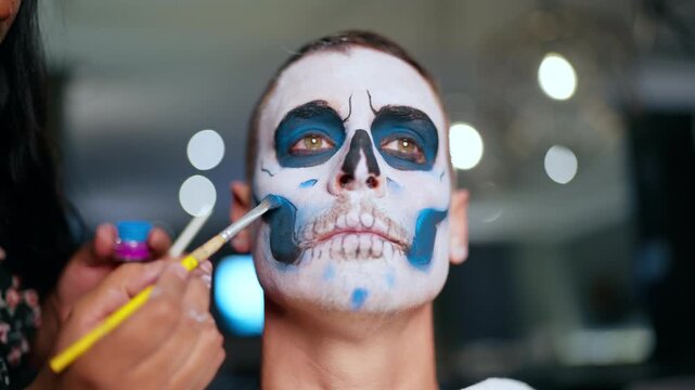 Professional makeup artist carefully applies traditional Day of the Dead or Halloween sugar skull face paint on young man using brush and blue paint with bokeh lights in background.