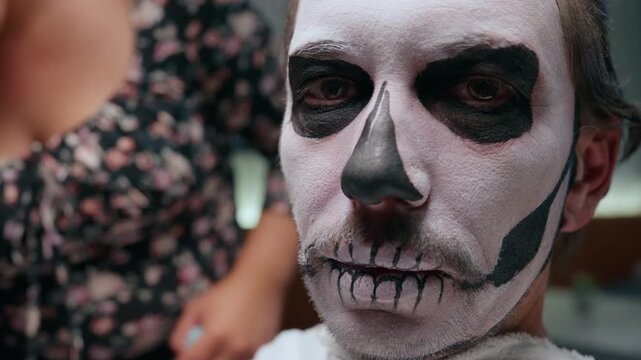 Close-up portrait of man with traditional Day of the Dead skull face paint featuring white base and black details around eyes and mouth. Makeup artist works in background.