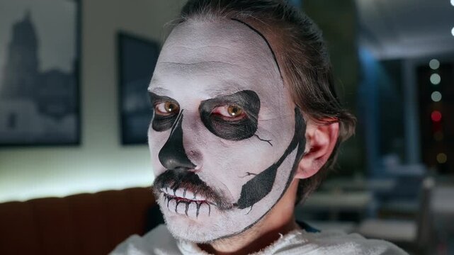Close-up portrait of man with detailed skull face paint makeup featuring black and white design around eyes nose and mouth for Day of the Dead or Halloween celebration indoors.
