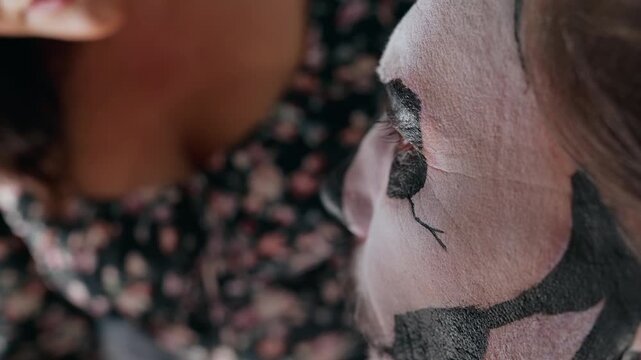 Close-up of makeup artist carefully painting black details on white face paint for Day of Dead or Halloween celebration. Skilled hands use fine brush to create traditional skull design around eye.