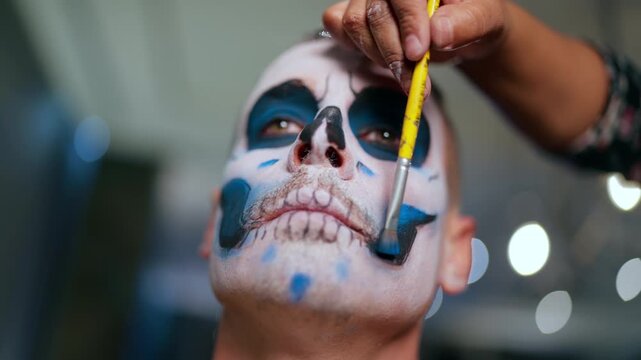 Makeup artist carefully applies intricate Day of the Dead sugar skull face paint on man using yellow brush. Close-up shows detailed white, black and blue design with bokeh lights in background.