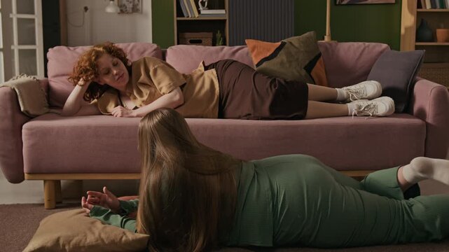 Caucasian young adult woman lying on sofa talking to curvy female friend lying on floor. Footage is ideal for body positivity campaigns and modern lifestyle advertisements