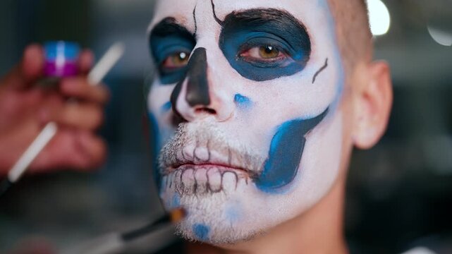 Close-up of makeup artist applying traditional Day of the Dead skull face paint on man. White base with intricate black and blue details around eyes and mouth creates dramatic calavera look.
