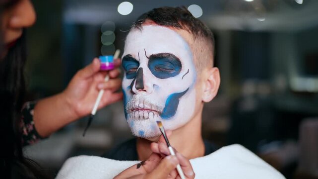 Professional makeup artist carefully applies Day of the Dead or Halloween skull face paint on young man using brushes and colorful pigments in studio setting with bokeh lights.
