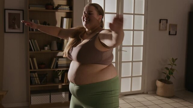 Plus-size Caucasian young adult woman exercising at home, smiling and performing arm swings. Clip is perfect for body positivity campaigns and home fitness marketing