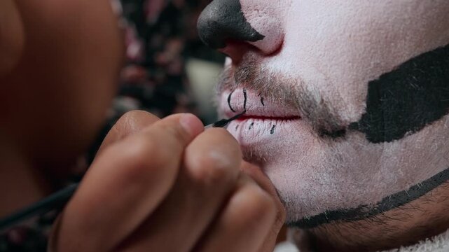 Makeup artist carefully applies traditional Day of the Dead skull face paint on man with white base and black details. Brush draws intricate patterns around mouth creating authentic catrina style.
