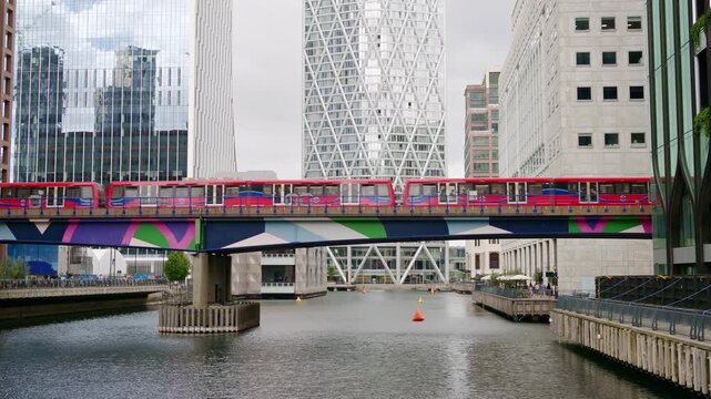 red train moving left to right over canal with modern city buildings as background on cloudy day.