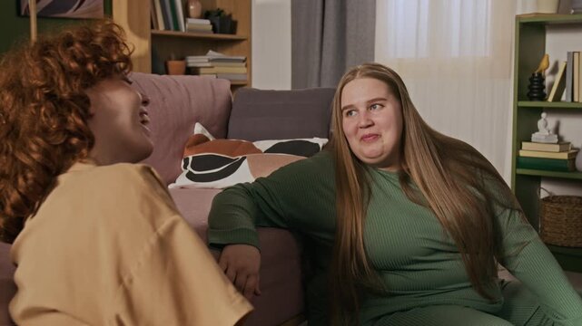 Two Caucasian young women, including plus-size female, sitting on floor by sofa, talking and laughing together. Clip is perfect for body positivity campaigns and lifestyle marketing