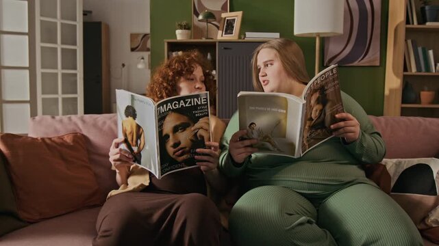 Two young adult Caucasian women, one plus-size, sitting on sofa, lowering fashion magazines from faces, talking, and laughing. Footage is perfect for body positivity and lifestyle marketing