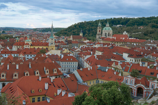 Panoramic high-angle view of Prague cityscape featuring traditional red tile roofs, historic architecture, and St. Nicholas Church under a cloudy sky.