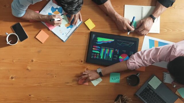 Top aerial view of diverse team writing colorful sticky notes to brainstorm marketing idea while looking at financial chart. Multicultural smart business people taking a note about plan. Convocation.