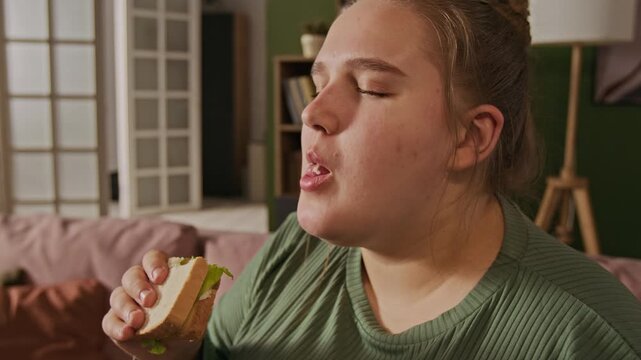 Plus-size Caucasian young adult woman eating sandwich, chewing and holding food while sitting on sofa indoors. Clip is suitable for body positivity concepts or fast food commercials