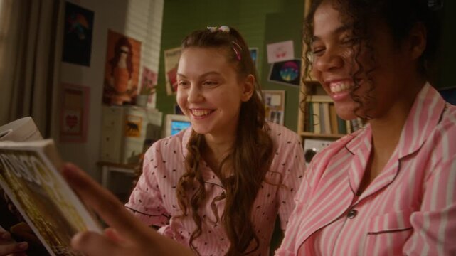 Caucasian teenage girl and African American friend reading fashion magazine during sleepover, smiling and pointing at pages. Perfect for youth lifestyle marketing and friendship campaigns