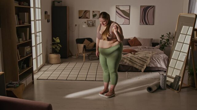 Plus-size Caucasian young adult woman stepping on scales in bedroom, smiling and celebrating weight loss progress. Perfect for body positivity campaigns and healthy lifestyle marketing