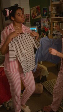 Vertical of African American female teenager and Caucasian female teenager in pajamas choosing outfits in front of mirror. Ideal for youth lifestyle and fashion retail