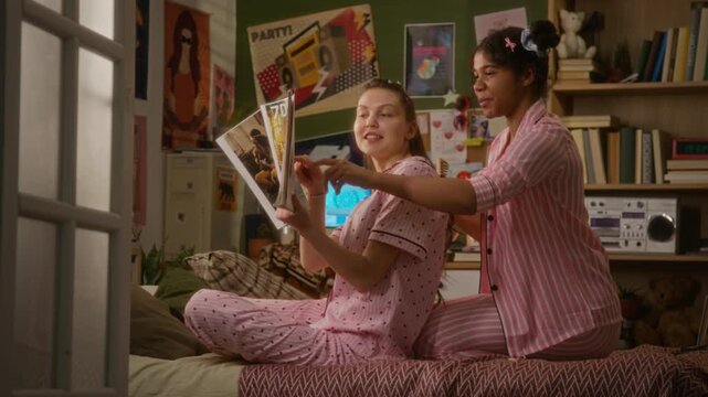 Black girl and Caucasian teenager sitting on bed in pajamas, reading magazine and brushing hair during sleepover. Ideal for youth lifestyle marketing and friendship campaigns