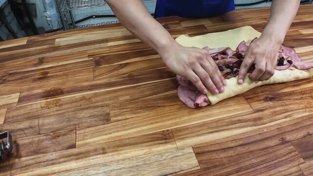 skilled artisan demonstrates the precise technique of rolling dough filled with savory ham, olives, and raisins in a commercial bakery setting.