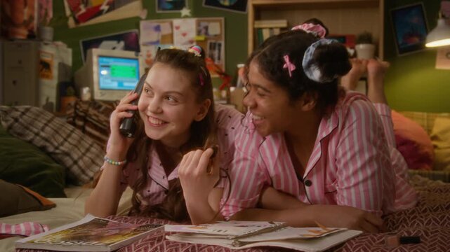 Caucasian teenager talking on retro cordless phone while lying on bed next to Black girlfriend during pajama party. Video suitable for nostalgic youth marketing and telecommunication ads