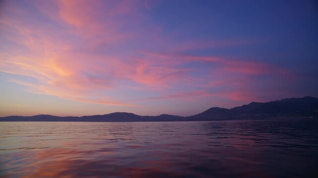 Peaceful sunset view from a boat on Lake Geneva, Switzerland. Pink clouds stretch across the sky above calm water and distant mountain silhouettes near the Jura.
