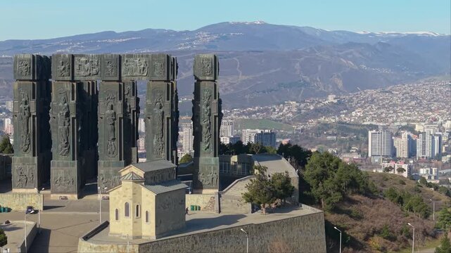 Lateral drone shot capturing the massive columns of the Chronicle of Georgia monument with Tbilisi stretching across the background.