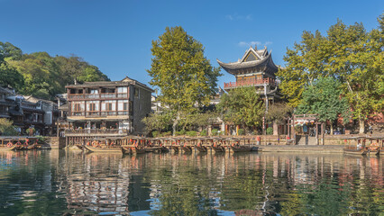 Fototapeta premium Tourist wooden boats with canopies and lifebuoys are moored in a row by the river bank. Reflection. Green trees. Chinese buildings with curved roofs against the blue sky. China. Fenghuang Ancient Town