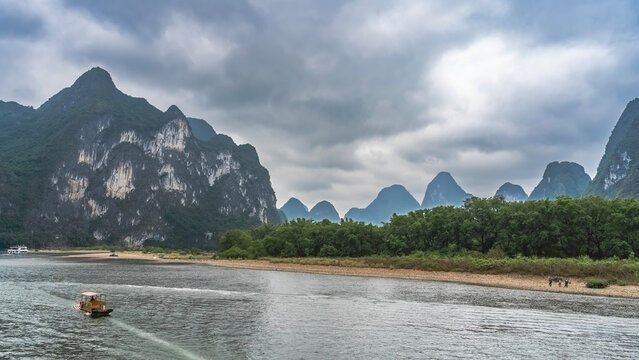 A bamboo tourist raft floats along a picturesque river. The ship is far away. There is green vegetation on the shore. Mountains against the sky and clouds. China. Li River. Li Jiang. Guilin