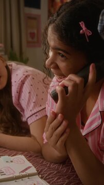 Vertical video of excited African American teenager talking on cordless phone while Caucasian girl friend listening on bed atr sleepover. Youth lifestyle and telecommunication marketing campaigns