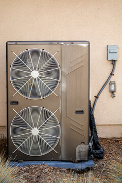 An outdoor heat pump or central air conditioner system with two large ventilation fans mounted against an exterior stucco wall