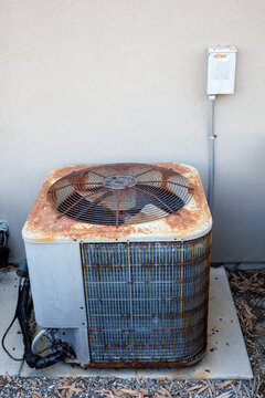 An old rusty outdoor central air conditioner condenser unit with a top ventilation fan installed next to a residential house wall