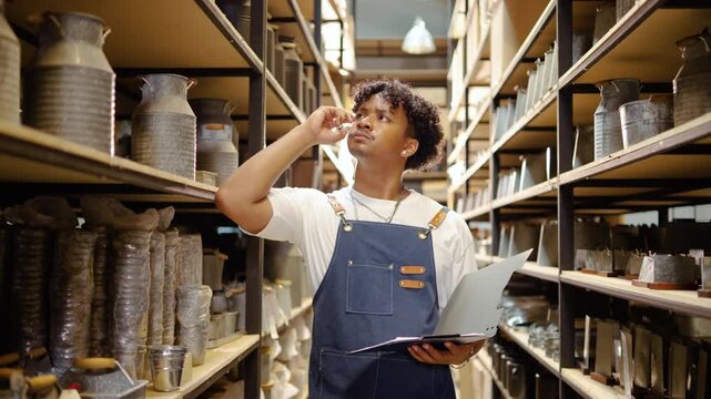 Focused young Black male artisan wearing an apron checks inventory in a rustic warehouse, holding a clipboard while inspecting metal pots on shelves with a thoughtful expression.