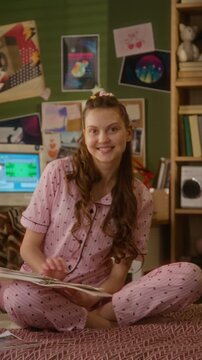 Portrait of Caucasian teenage girl sitting on bed in pajamas, looking at scrapbook journal and smiling at camera. Youth lifestyle, hobby, or creative education marketing campaigns, vertical