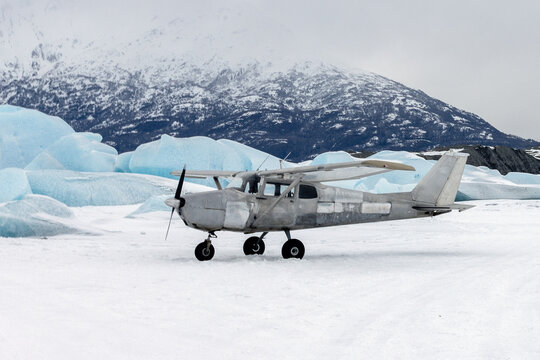An unpainted aluminum Cessna light aircraft parked on the snowy surface of Knik Glacier with the rugged Chugach Mountains of Alaska in the background.