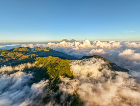 Aerial view of the lush green slopes of Gunung Bismo rising above a sea of clouds during golden hour near the basecamp in Kecamatan Kejajar, Jawa Tengah, Indonesia.