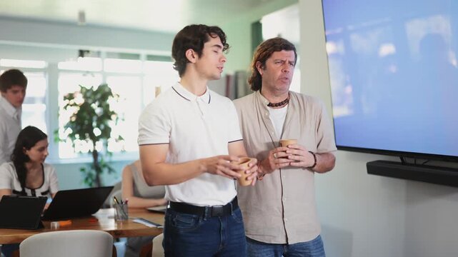 Colleagues share positive thoughts during coffee break. Two guys talking, chatting and joking with cup of coffee in hands, modern working atmosphere in office 