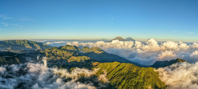 Aerial view of the mountainous landscape near Basecamp Gunung Bismo, with green hills, rolling clouds, and distant volcanic peaks in Kecamatan Kejajar, Jawa Tengah, Indonesia.
