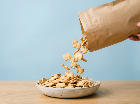 Animal cracker cookies falling from paper bag, crunchy biscuit snacks pouring into a bowl, sweet treats for kids on blue background