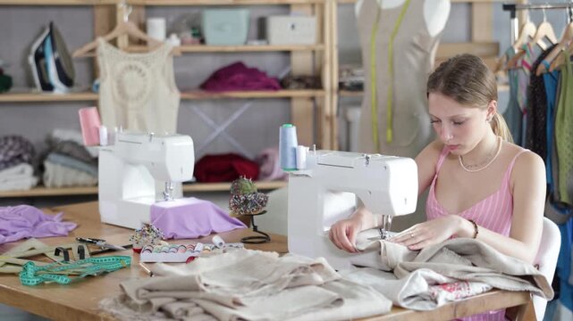 Young dressmaker sitting and sewing fabric on a sewing machine. Seamstress in the workshop. Process of tailoring clothes according to individual measurements and sketches