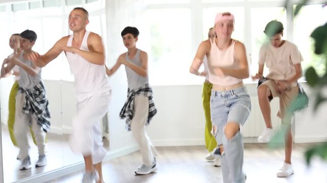 Girl in baseball cap with two hair pigtails with group of teen friends and male teacher, learn to dance hip hop in choreographic hall. Students moving, do moonwalking, squats, bends