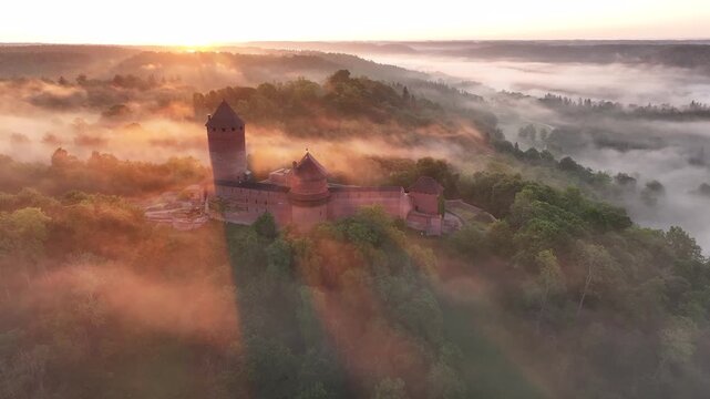 Beautiful aerial drone footage of Turaida Castle rising above the morning mist in Gauja National Park near Sigulda, Latvia. Warm sunrise light illuminates the medieval red brick fortress.