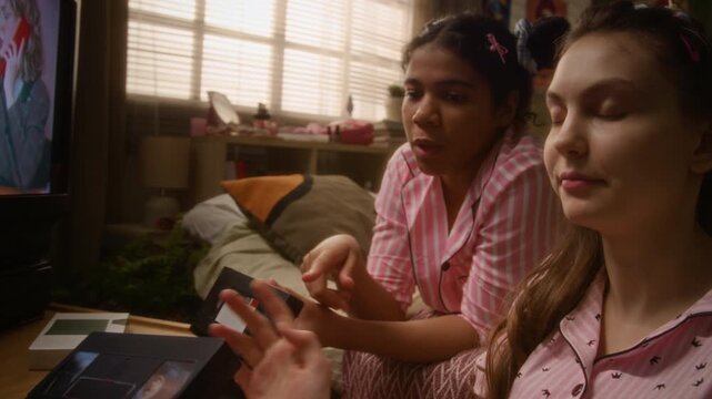 Diverse teenage grilfriends, Caucasian and African American, holding VHS tapes and talking while choosing movie during slumber party with retro TV playing. Nostalgia marketing, youth lifestyle