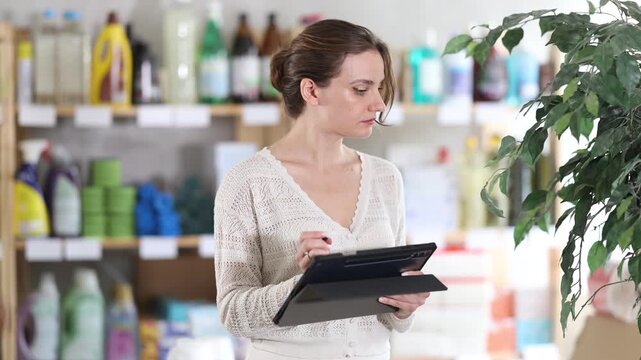  Young woman seller in uniform conducting inventory with tablet in household chemicals store