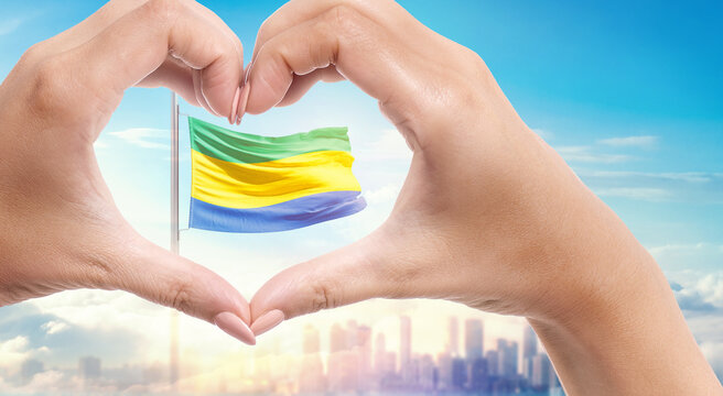 Gabon national flag. Two hands forming a heart shape in front of a national flag waving in beautiful clouds. Gabon national flag behind the heart shape hand.