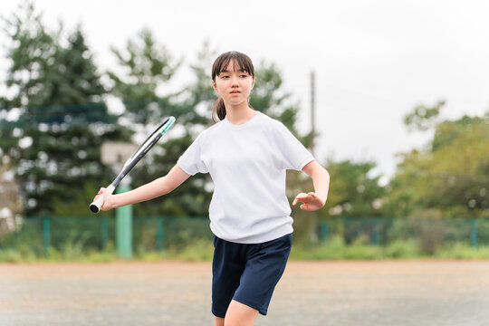 Elementary, middle, and high school girls in gym clothes playing tennis on the school grounds.