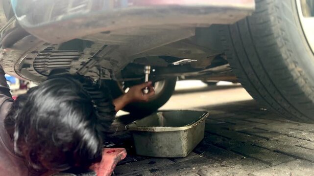 A mechanic lies beneath a car on a wheeled creeper, changing engine oil in a workshop. Rear view with no visible face, illustrating manual labor, automotive maintenance, and repair service.