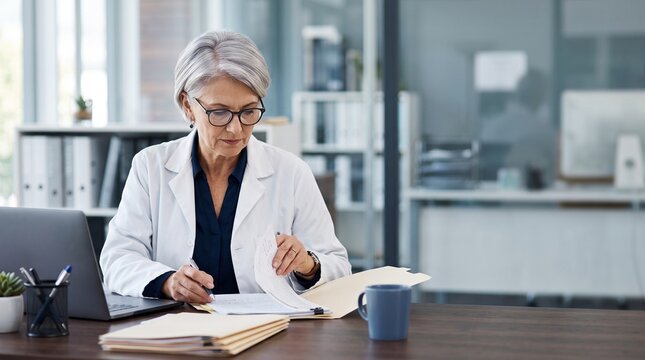 Mature senior woman doctor physician in white medical uniform reviewing patient medical history records documentation file at desk with laptop in hospital office clinical concept