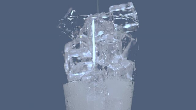 A close-up of ice cubes stacked in a glass with soda being poured creating splashes against a gray background.