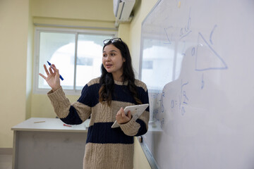 A dynamic young female educator holds a digital tablet and marker as she actively explains a geometry and mathematics lesson on a whiteboard in a modern classroom setting. © Nalinee