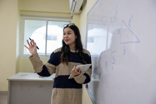 A dynamic young female educator holds a digital tablet and marker as she actively explains a geometry and mathematics lesson on a whiteboard in a modern classroom setting.