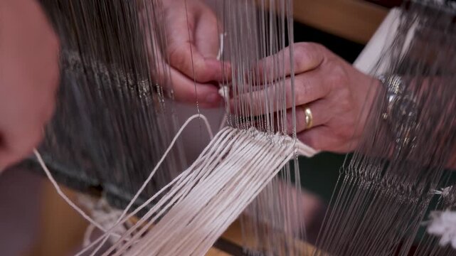 Detailed close-up of an experienced textile artisan carefully threading white warp yarns through metal heddles on a traditional handloom. The precise preparation stage is essential for creating handma